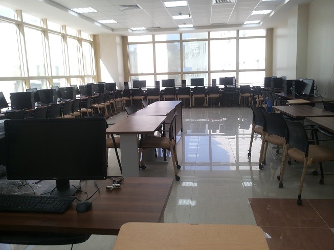 A spacious computer lab with multiple desks and chairs, featuring several desktop computers and large windows for natural light.